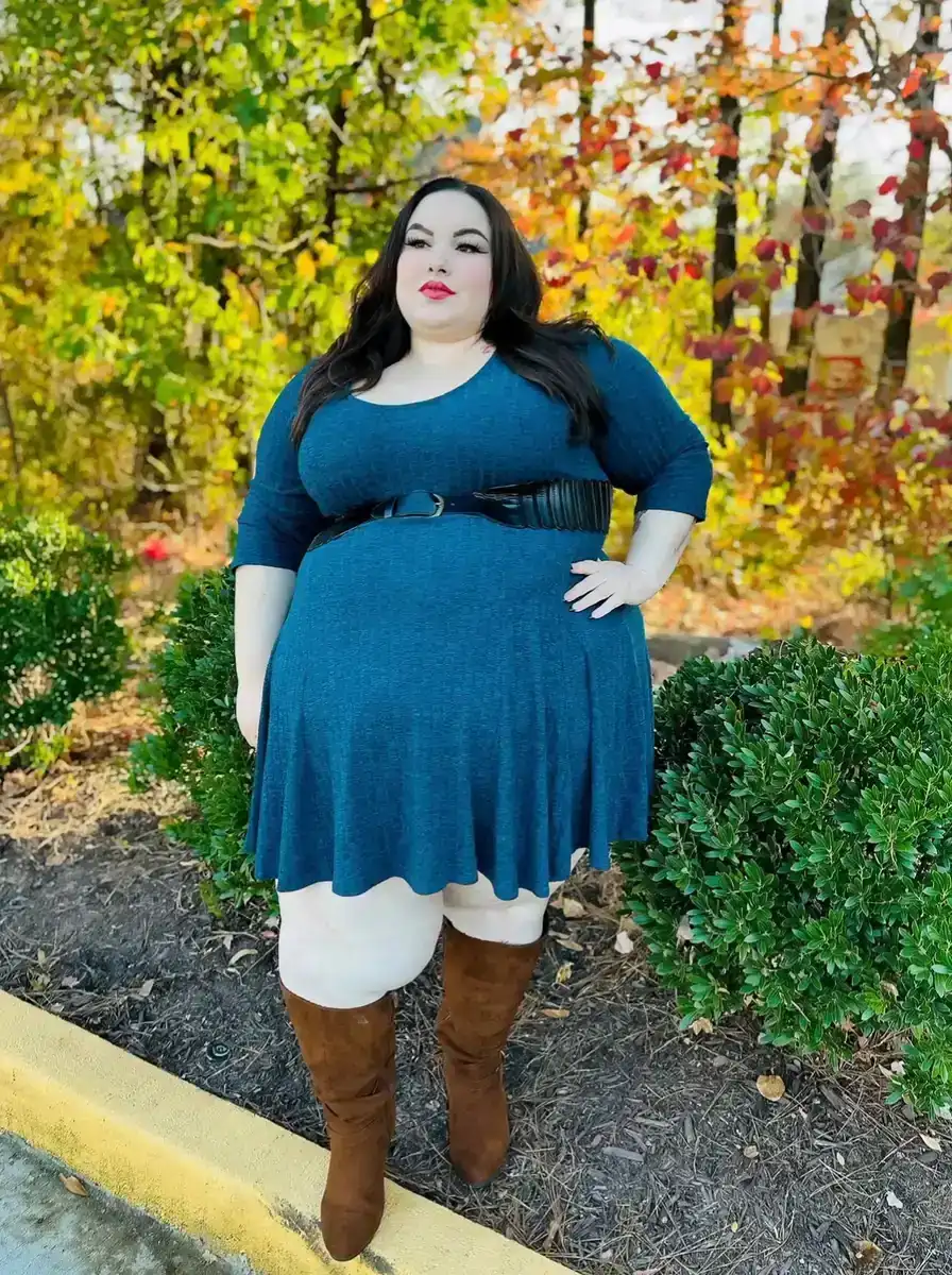 Plus size woman wearing a teal belted sweater dress with white tights and brown knee-high boots in autumn