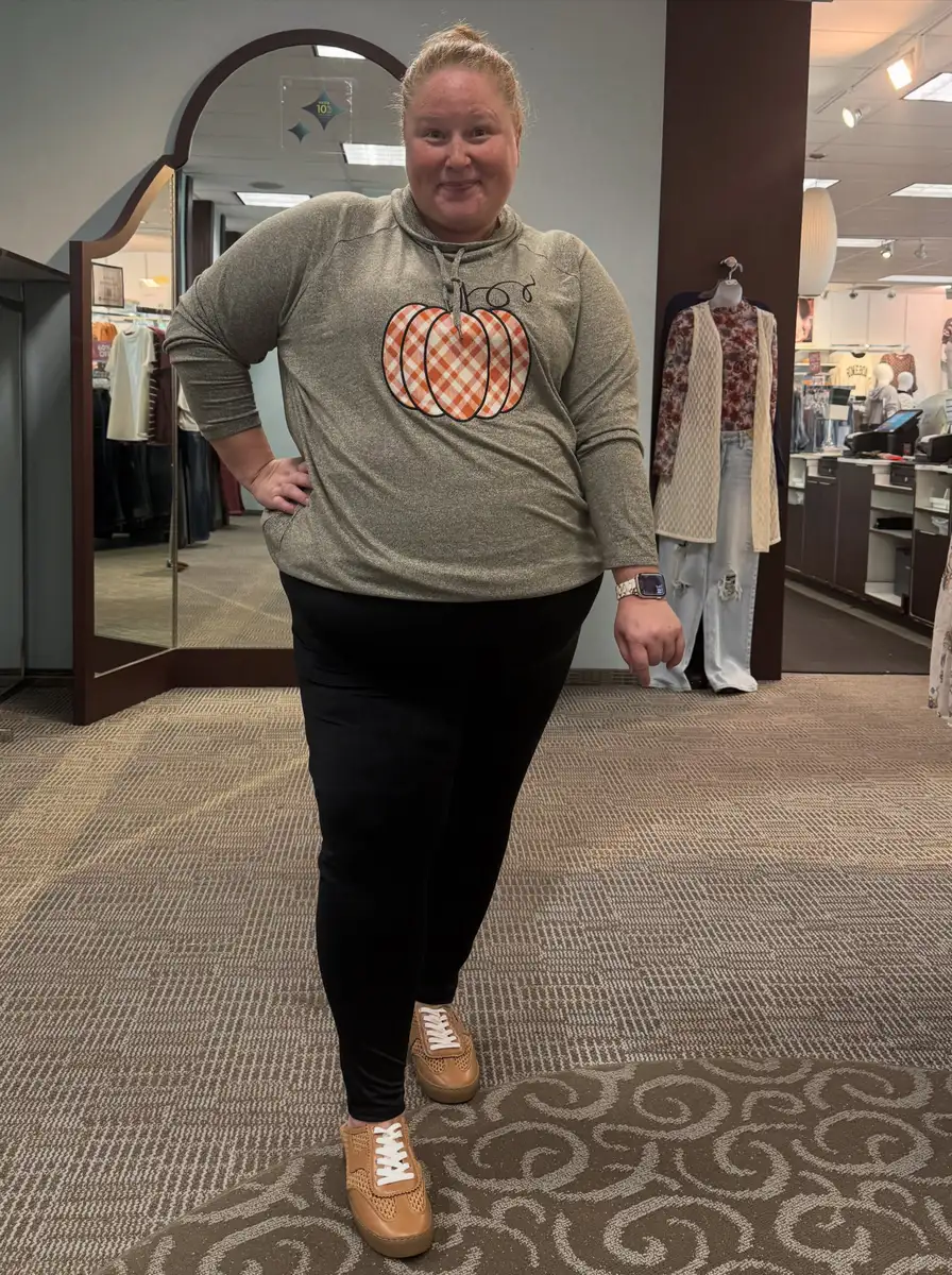 Plus size woman wearing a pumpkin graphic cowl-neck top with black leggings and tan sneakers