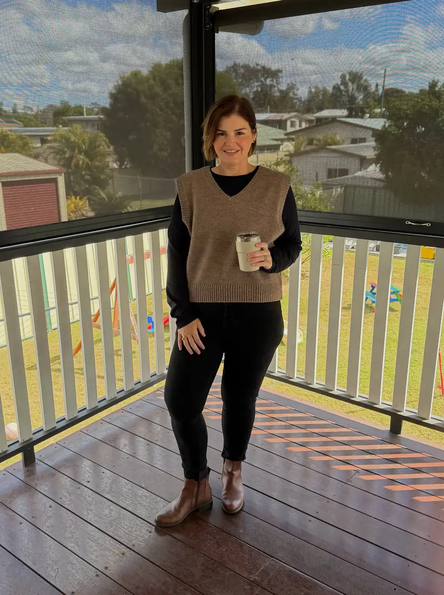Plus size woman wearing a taupe knit sweater vest over a black top with black jeans and tan boots