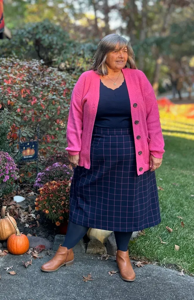 Older plus-size woman wearing a hot-pink cardigan over a navy plaid midi skirt in a garden with pumpkins