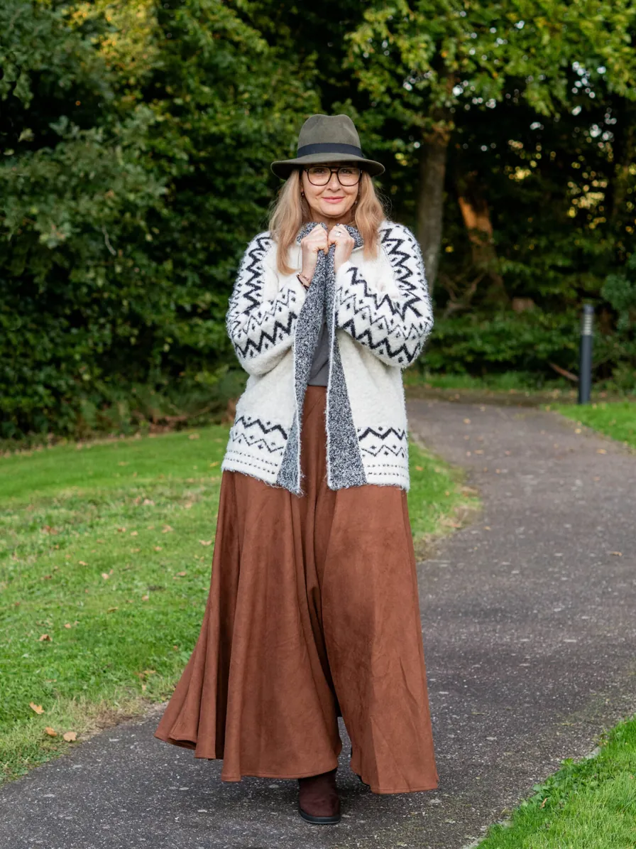 Plus-size woman over 50 in a cream fair-isle cardigan, rust wide-leg pants, and a gray fedora on a park path