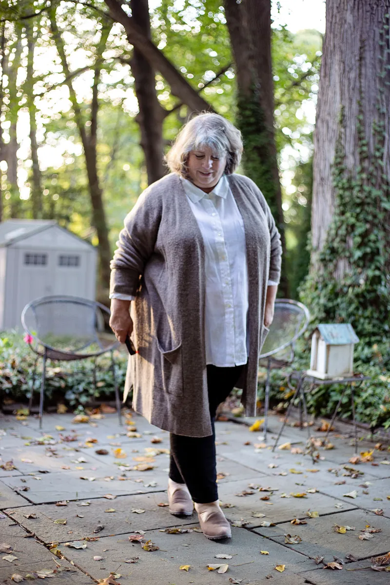 Plus-size woman over 50 wearing a taupe duster cardigan over a white shirt with black pants among fall leaves