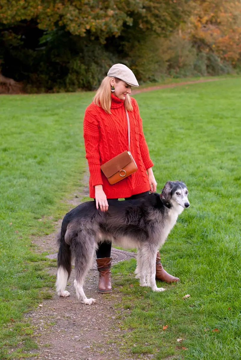 Woman in a red cable-knit turtleneck and black leggings standing with a large dog on grass