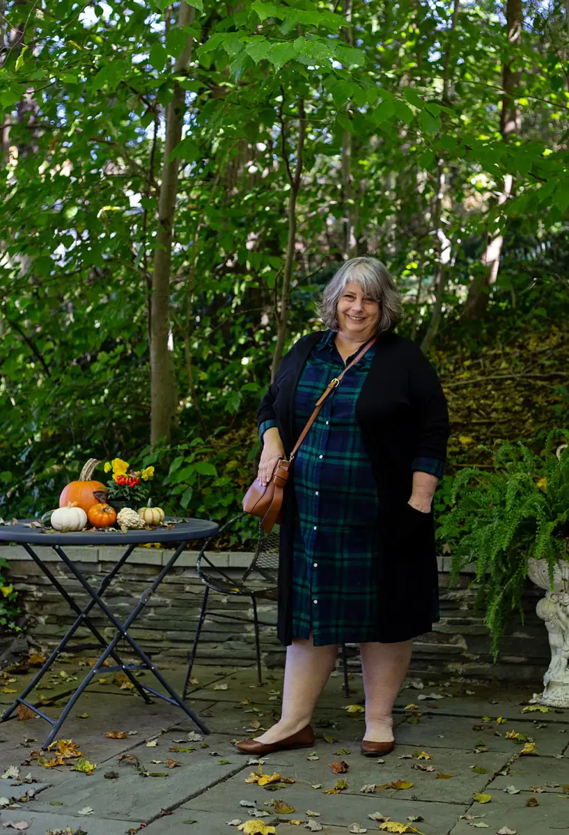Plus-size woman over 50 in a green tartan shirtdress with a black duster cardigan on a patio with pumpkins