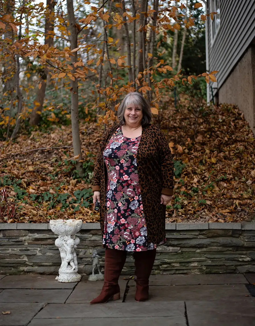 Mature plus-size woman in a floral midi dress, leopard cardigan, and rust knee-high boots among autumn leaves
