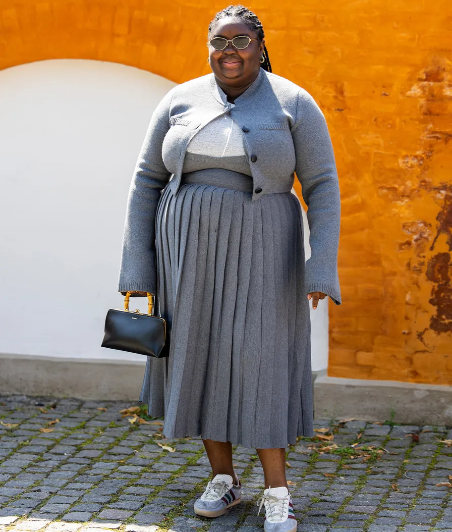 Plus-size woman in a grey monochrome knit cardigan, top, and pleated midi skirt with white sneakers