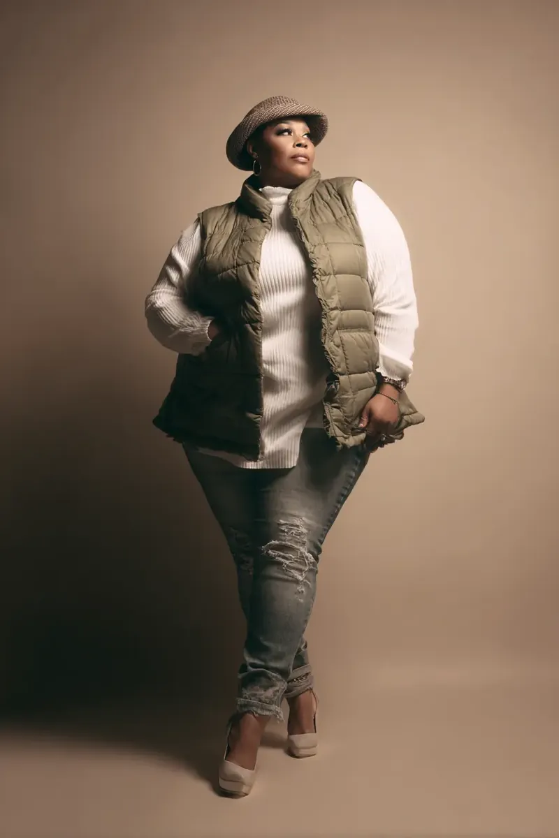 Plus-size woman in an olive quilted puffer vest over a white turtleneck with ripped jeans, nude heels, and a tweed flat cap